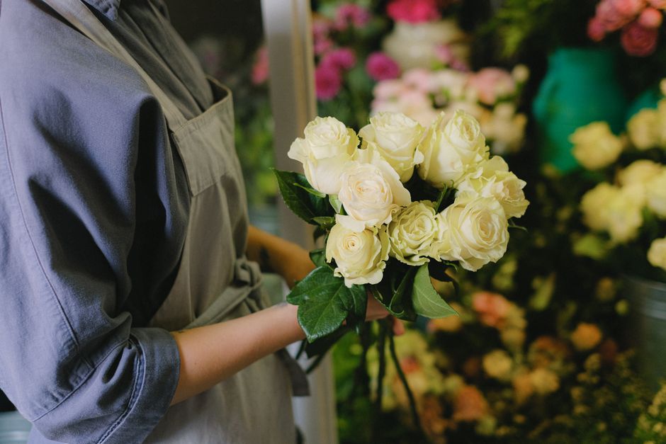 A person holding a bouquet of fresh, cream-colored roses with lush green leaves, wrapped loosely in clear and white paper, standing in a floral shop. The arrangement is compact and symmetrical, showca