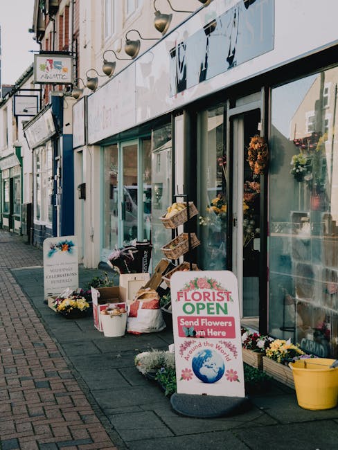 A storefront of a flower shop on Wallington High Street displays a variety of floral arrangements and supplies. Outside, there are buckets and boxes filled with fresh flowers, including chrysanthemums